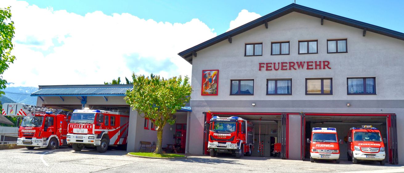 Eine Feuerwehrstation mit einem roten Feuerwehrauto vor dem Gebäude. Das Gebäude hat ein rotes Schild mit dem Wort 'FEUE' und einem Bilderrahmen. Vor dem Gebäude steht ein Baum.