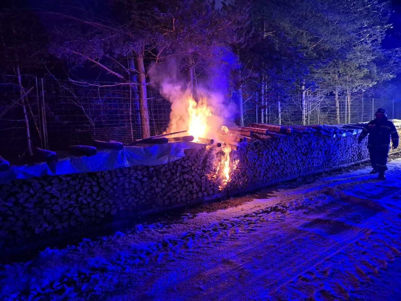 Eine nächtliche Szene mit einem Feuer, das durch einen Holzstapel in einer verschneiten Landschaft brennt, beleuchtet von blauen Lichtern.