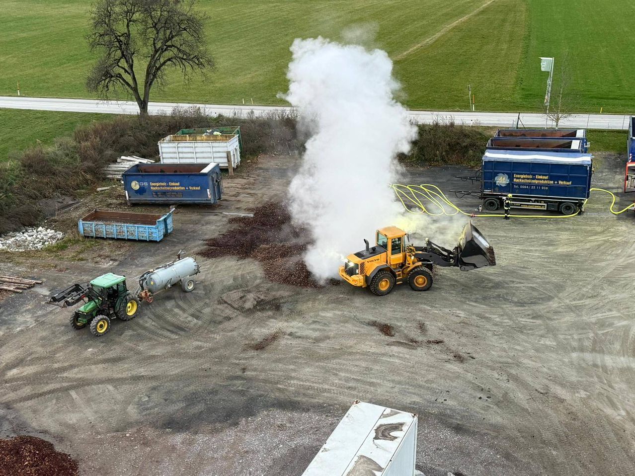 An aerial view of a construction site with heavy machinery. A yellow tractor is dumping dirt, and white smoke is coming from the area. Nearby, there are several containers and a truck.