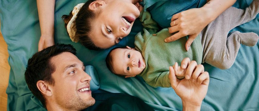Eine lächelnde Familie mit einem Baby liegt auf einem Bett, die Eltern schauen ihr Kind an, das die Hand ausstreckt.