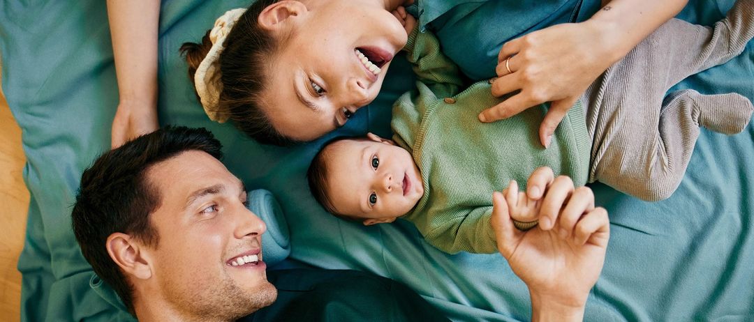 Eine lächelnde Familie mit einem Baby liegt auf einem Bett, die Eltern schauen ihr Kind an, das die Hand ausstreckt.