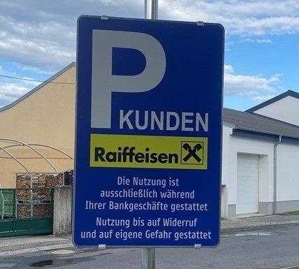 Blaues Schild mit weißem Buchstaben P. Darunter steht 'Raiffeisen Kunden'. Gelbes Kreuz neben 'Raiffeisen'. Darunter steht 'Die Nutzung ist ausschließlich während Ihrer Bankgeschäfte gestattet'. Darunter steht 'Nutzung bis auf Widerruf und auf eigene Gefahr gestattet'. Hinter dem Schild ist ein Haus zu sehen.