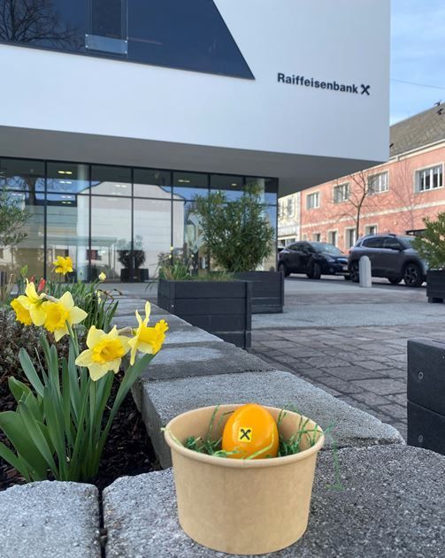 A small bowl with an orange egg is placed on the ground in front of the Raiffeisenbank building. The area is decorated with yellow flowers and plants.