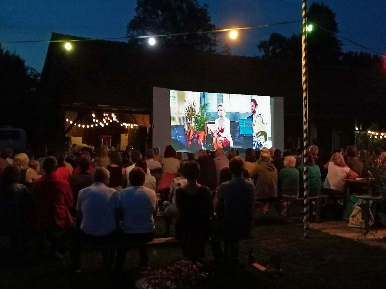 A group of people are watching a movie on a large screen outdoors at night. The screen shows a man and a woman.