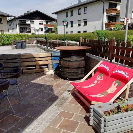 Ein Patio mit einem Holzfass und einem roten Liegestuhl mit Strandzeichnungen. Stühle sind herum platziert. Ein Holztisch steht in der Nähe des Fasses. Gebäude befinden sich hinter dem Zaun.