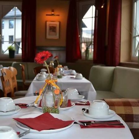 Ein Restaurant mit mehreren Tischen, die jeweils mit Tellern, Tassen und Servietten besetzt sind. Auf den Tischen befinden sich ein Blumenvase, Salz- und Pfefferstreuer. Der Raum hat große Fenster mit roten Vorhängen und Topfpflanzen auf den Fensterbänken.