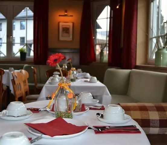 Ein Restaurant mit mehreren Tischen, die jeweils mit Tellern, Tassen und Servietten besetzt sind. Auf den Tischen befinden sich ein Blumenvase, Salz- und Pfefferstreuer. Der Raum hat große Fenster mit roten Vorhängen und Topfpflanzen auf den Fensterbänken.