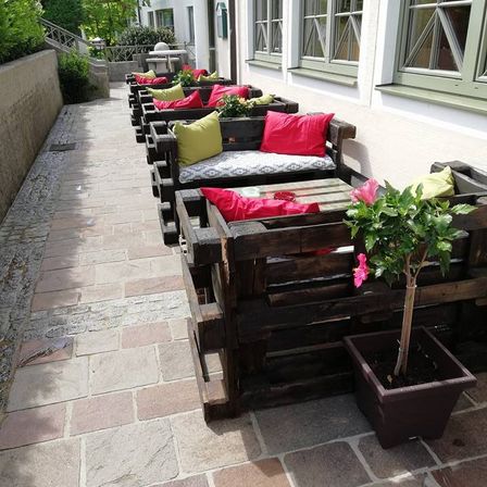 Ein Außensitzbereich mit Holzbänken und bunten Kissen, begleitet von einer kleinen Topfpflanze. Der Bereich ist mit Ziegelsteinen gepflastert, mit Fenstern und einer Wand im Hintergrund.