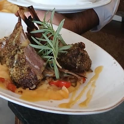 Ein Teller mit Lamm mit Rosmarin-Garnierung, serviert mit Kartoffelpüree und roten Paprikaschoten, begleitet von einem Saucen-Träufelchen, gehalten von einem Kellner.