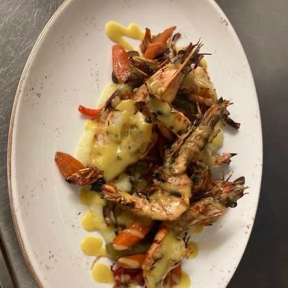 Ein weißer Teller mit gegrillten Garnelen, Karotten und Paprika, bedeckt mit einer gelben Sauce, auf einer Metalloberfläche platziert.