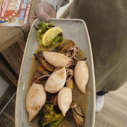 Eine Person hält einen Teller mit Meeresfrüchten, bestehend aus Tintenfisch, Brokkoli und Pilzen, begleitet von einer Zitronenscheibe und einem Salat, vor einem hölzernen Hintergrund.