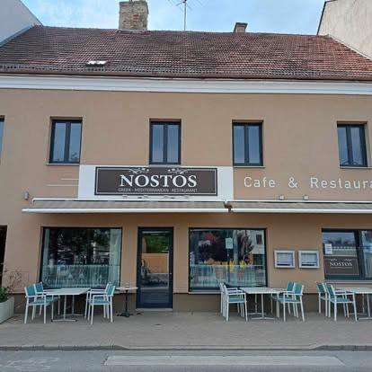 Die Vorderansicht eines Restaurants namens 'Nostos' mit einem Schild über dem Eingang. Es hat ein braunes Dach, Glasfenster und mehrere Tische und Stühle, die draußen aufgestellt sind.