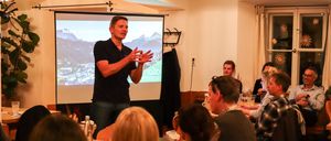 Ein Mann in einem schwarzen T-Shirt hält eine Präsentation für eine Gruppe von Menschen. Die Präsentation wird auf einer Leinwand hinter ihm projiziert, und es sind Berge auf dem Bildschirm sichtbar.