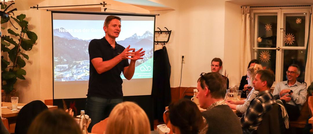 Ein Mann in einem schwarzen T-Shirt hält eine Präsentation für eine Gruppe von Menschen. Die Präsentation wird auf einer Leinwand hinter ihm projiziert, und es sind Berge auf dem Bildschirm sichtbar.