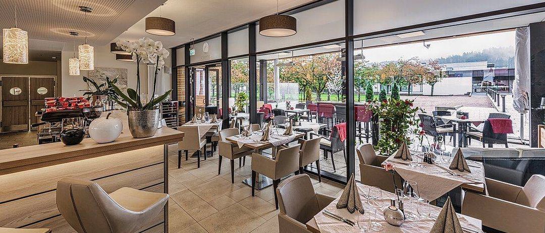 Ein Innenrestaurant mit moderner Einrichtung, weißen Blumen und großen Fenstern mit Blick auf das Freie.