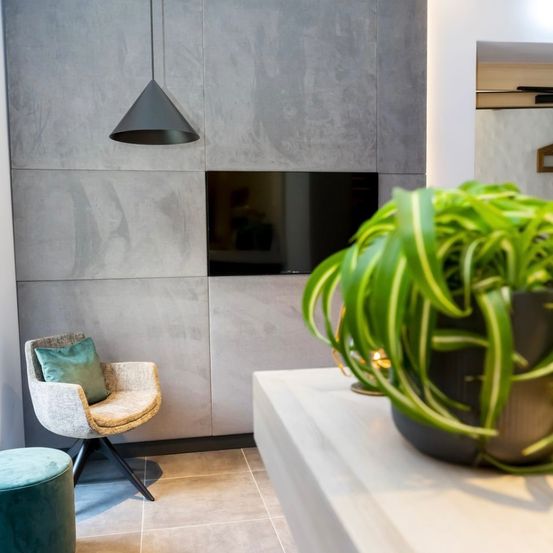 Ein moderner Raum mit grauen Wänden, einem Stuhl mit Kissen, einem an der Wand montierten Fernseher, einer Pendelleuchte, einer Topfpflanze und einem Hocker.