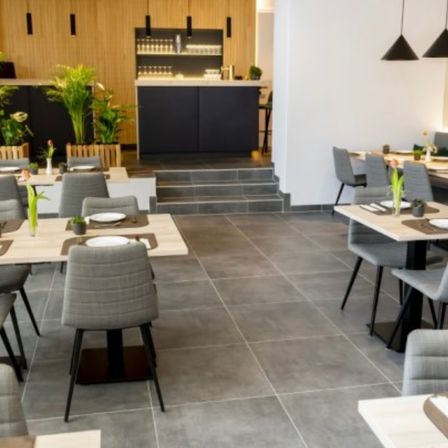 Ein modernes Restaurant-Interieur mit Holzboden, weißen Wänden und einer Theke. Mehrere Tische und Stühle sind aufgestellt, jeder mit einem Teller, Besteck und Blumen. Es gibt Topfpflanzen und Lampen.