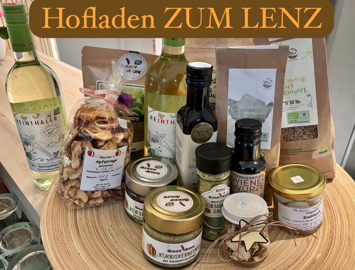 Ein Korb mit verschiedenen Produkten wie getrockneten Äpfeln, Nüssen und Flaschen mit Wein, Öl und Essig steht auf einem Holztisch. Die Flaschen haben Etiketten wie 'Reinth', 'BIOH' und 'Wiener Wurze'. Säcke mit Kräutern und Gewürzen sind ebenfalls vorhanden.