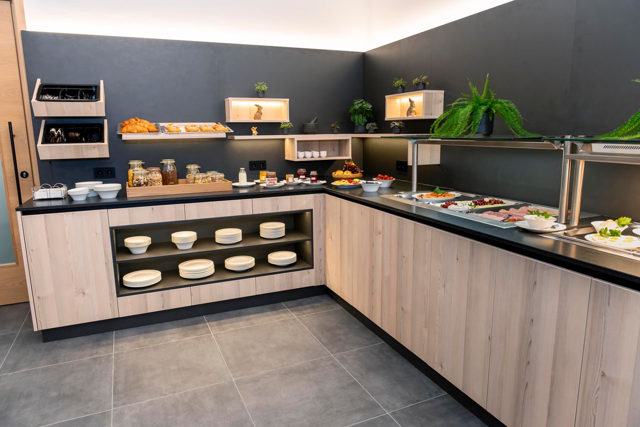 Eine moderne Küchenzeile mit grau gefliestem Boden und Holzschränken. Es gibt ein Frühstücksbuffet mit Brot, Obst und Desserts auf der Arbeitsplatte.