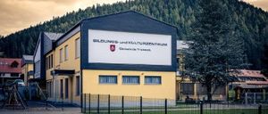 Ein großes gelbes Gebäude mit schwarzem Dach zeigt ein großes Schild mit der Aufschrift 'Bildungs- und Kulturzentrum'. Es ist von einem Zaun und einem Rasen umgeben. Im Hintergrund befinden sich Berge.