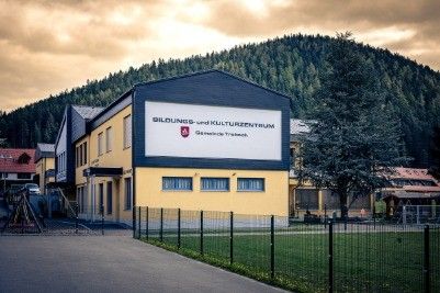Ein großes gelbes Gebäude mit schwarzem Dach zeigt ein großes Schild mit der Aufschrift 'Bildungs- und Kulturzentrum'. Es ist von einem Zaun und einem Rasen umgeben. Im Hintergrund befinden sich Berge.