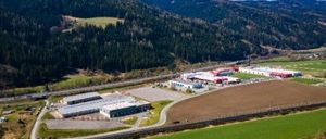 Luftaufnahme eines Industriegebiets mit mehreren Gebäuden und einem weitläufigen grünen Feld, eingebettet zwischen Bergen und unter einem klaren blauen Himmel.