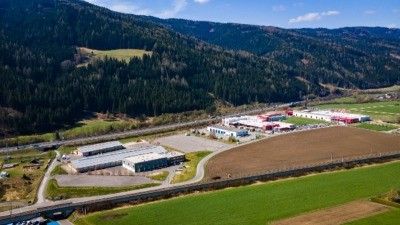 Luftaufnahme eines Industriegebiets mit mehreren Gebäuden und einem weitläufigen grünen Feld, eingebettet zwischen Bergen und unter einem klaren blauen Himmel.