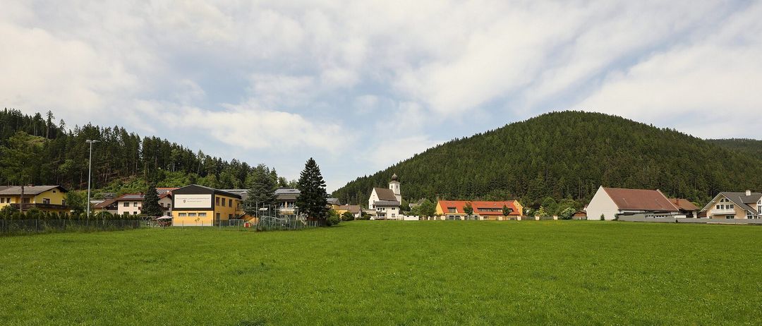Eine ländliche Szene mit einem großen grünen Feld und mehreren Häusern im Hintergrund. Die Häuser haben eine gelbe und weiße Außenseite. In der Mitte befindet sich eine Kirche mit einem Turm. Der Himmel ist bewölkt.