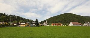 Eine ländliche Szene mit einem großen grünen Feld und mehreren Häusern im Hintergrund. Die Häuser haben eine gelbe und weiße Außenseite. In der Mitte befindet sich eine Kirche mit einem Turm. Der Himmel ist bewölkt.