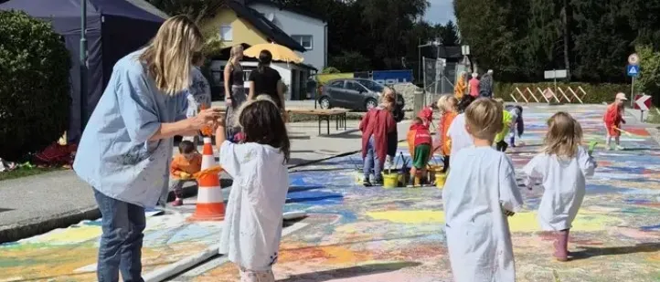 Eine Gruppe von Kindern und einem Erwachsenen malt ein großes Wandbild auf der Straße. Sie verwenden Farbbecher und Pinsel. Die Szene ist im Freien mit Bäumen und einem Haus im Hintergrund.