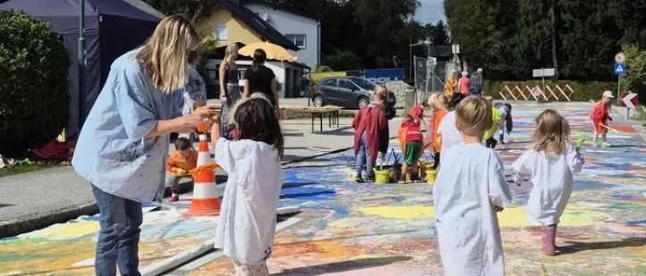 Eine Gruppe von Kindern und einem Erwachsenen malt ein großes Wandbild auf der Straße. Sie verwenden Farbbecher und Pinsel. Die Szene ist im Freien mit Bäumen und einem Haus im Hintergrund.