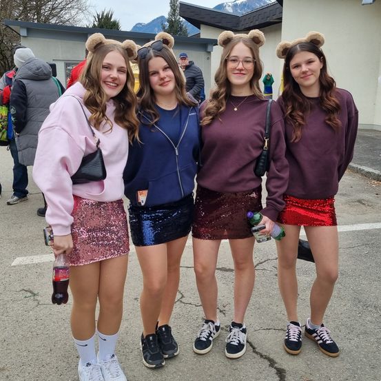 Vier junge Frauen mit Bärenohr-Kopfbändern posieren für ein Foto draußen, Getränke in der Hand, mit einem Gebäude und einem Berg im Hintergrund.