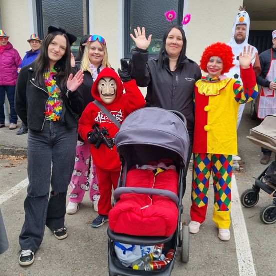 Eine Gruppe von Menschen in verschiedenen Kostümen steht draußen. Ein Clown, eine Frau und ein maskierter Mann stehen vorne, mit anderen dahinter. Ein Kinderwagen steht in der Mitte.