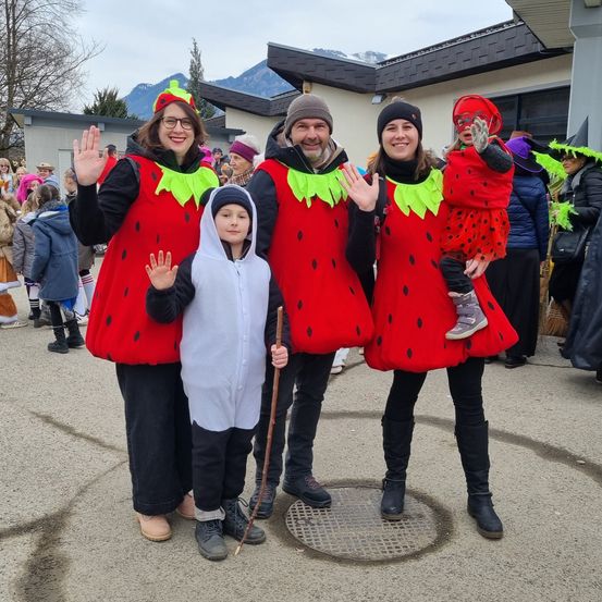 Eine Familie in Erdbeerkostümen steht mit einem Kind in einem grauen Hoodie. Dahinter sind Menschen in verschiedenen Kostümen gekleidet, mit einem Gebäude und Bergen im Hintergrund.