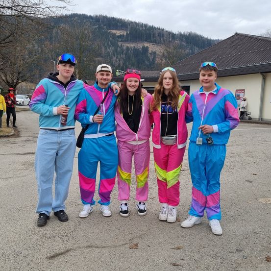 Fünf junge Leute in leuchtenden Overalls, zwei Mädchen und drei Jungen, posieren zusammen vor einem Gebäude mit Bergkulisse.