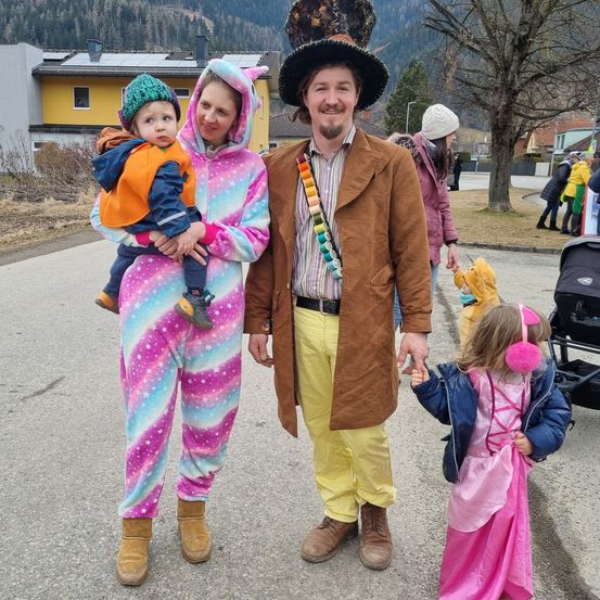 Eine Familie in Kostümen, wobei ein Erwachsener ein Baby hält und ein anderer Erwachsener die Hand eines Kindes hält, in einer Außenumgebung.