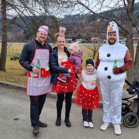Eine Familie in Kostümen steht auf einer Straße mit einem Kinderwagen dahinter. Ein Mann in einer Kochmütze, eine Frau in einem Popcorn-Kostüm und ein Mädchen in einer roten Tutu. Ein Mann in einem Schneemann-Kostüm und ein Baby in einer rosa Mütze. Bäume und eine Wiese sind im Hintergrund.