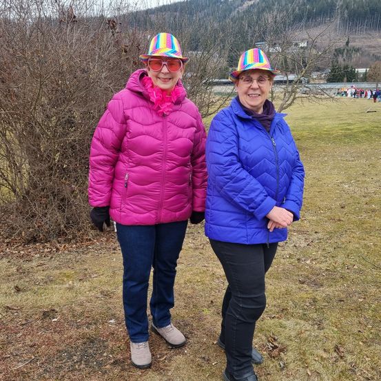Zwei Frauen stehen in einem Feld mit einem Berg im Hintergrund, eine trägt eine rosa Jacke und einen Regenbogenhut, die andere eine blaue Jacke und einen bunten Hut.
