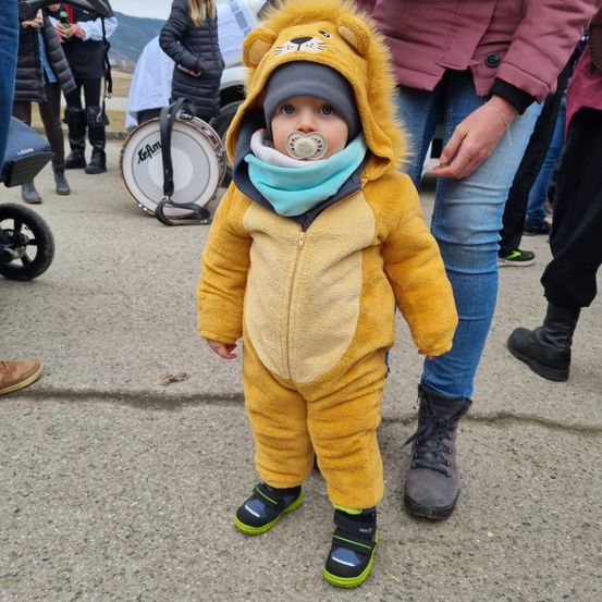 Ein Baby in einem Löwenkostüm mit einem Schnuller, steht draußen mit einem Erwachsenen in der Nähe. Im Hintergrund sind mehrere Leute, einige tragen Stiefel und tragen eine Trommel.
