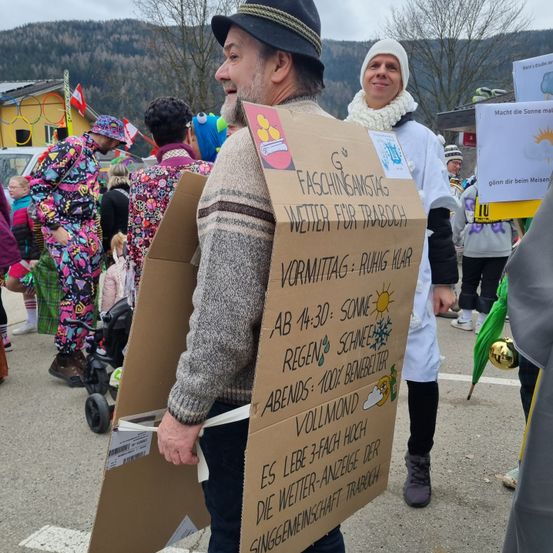 Ein Mann mit einem Schild mit Wettervorhersagen für Faschingstag steht in einer Menschenmenge bei einer Veranstaltung im Freien. Hinter ihm lächelt eine Person in einem weißen Mantel. Im Hintergrund sind ein Berg, Bäume und ein Gebäude zu sehen.