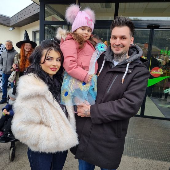 Eine Familie posiert für ein Foto vor einem Geschäft, mit einer Frau in Pelz, einem Mann in einer Kapuzenjacke und einem jungen Mädchen in einem rosa Mantel.