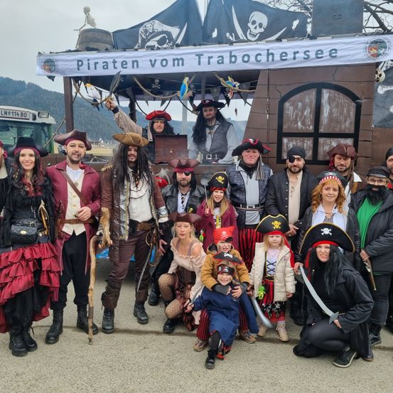 Eine Gruppe von Menschen, die als Piraten verkleidet sind, posiert für ein Foto vor einem Piratenschiff. Sie alle lächeln und halten Requisiten.