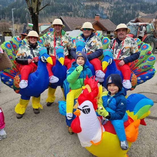 Eine Gruppe von Menschen in aufblasbaren Vogelkostümen, mit Erwachsenen und einem Kind, posiert für ein Foto bei einem festlichen Event. Berge sind im Hintergrund zu sehen.