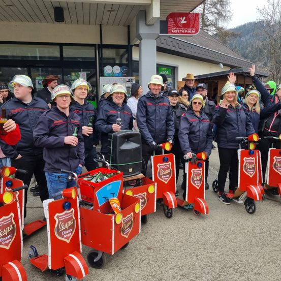 Eine Gruppe von Menschen in gleichen Jacken posiert mit roten Rollern vor einem Geschäft mit einem roten Schild. Sie lächeln und halten Flaschen.