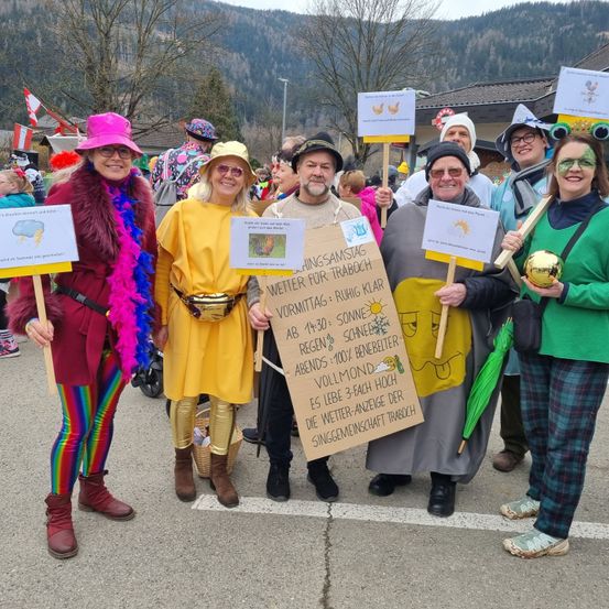 Eine Gruppe von Erwachsenen in Kostümen, einer hält ein Schild mit der Aufschrift 'WETTERSAMSTAAG: RUHIG KLAR VORMITAAGS: SONNE AB 14:30 SCHNEE 100% BENEITEER ABENDS VOLLEMOND' und eine andere hält eine Kugel.
