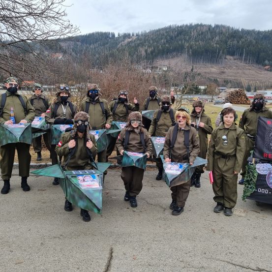 Eine Gruppe von Menschen in Militäruniformen posiert draußen mit grünen Objekten, die Flugzeuge ähneln. Sie tragen Masken, Schutzbrillen und Mützen, und ein Banner ist hinter ihnen. Im Hintergrund sind ein Berg und Bäume zu sehen.