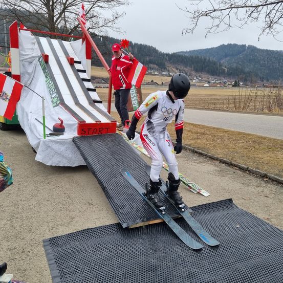 Eine Person fährt mit Skiern eine Rampe hinunter, hinter ihr weht eine Flagge. Die Rampe hat ein Schild, das Start sagt. Eine Flagge fliegt über der Rampe.