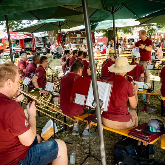 Eine Band in roten T-Shirts spielt Instrumente unter grünen Schirmen bei einer Outdoor-Veranstaltung. Zuschauer schauen von Picknick-Tischen zu. Ein Mann steht und gestikuliert.