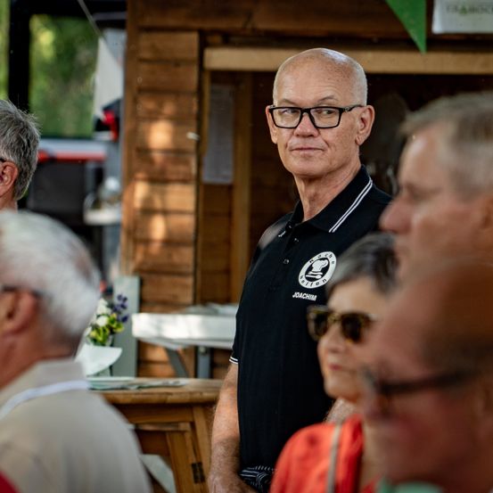 Ein Mann mit Brille und schwarzem Poloshirt steht vor einer Gruppe von Menschen, wahrscheinlich bei einer Veranstaltung im Freien.