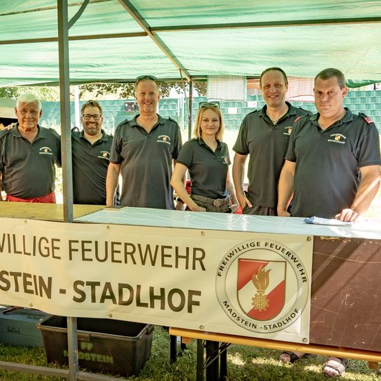 Fünf Personen in Uniform stehen hinter einem Tisch mit einem Banner, das 'Freiwillige Feuerwehr Madstein-Stadtlhof' besagt. Sie lächeln für die Kamera.